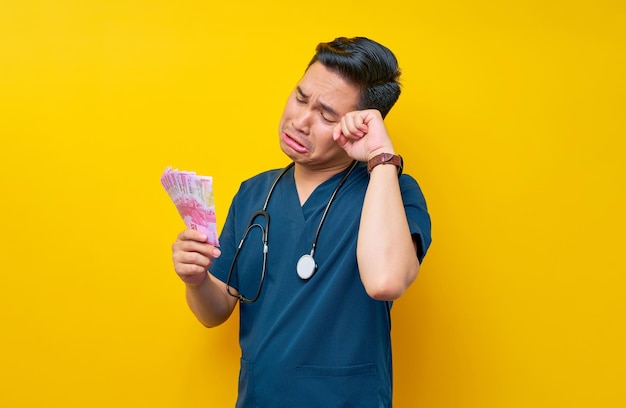 Foto porträt eines unglücklichen jungen asiatischen männlichen arztes oder krankenschwestern in blauer uniform mit bargeld isoliert auf gelbem hintergrund gesundheitsmedizin konzept