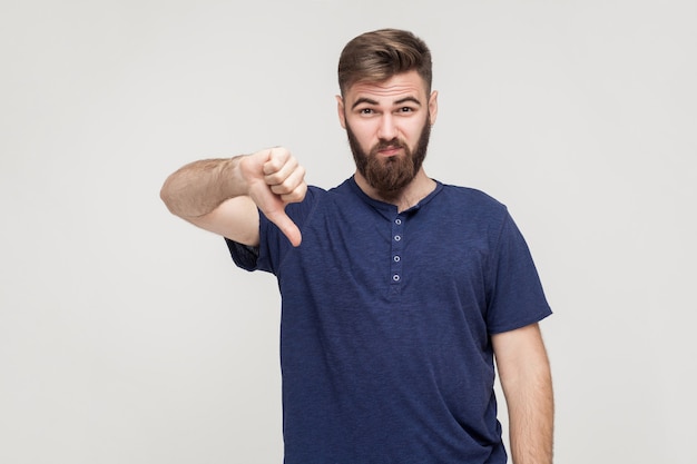 Porträt eines unzufriedenen bärtigen Mannes mit Daumen nach unten und blauem T-Shirt vor grauem Hintergrund. Studioaufnahme.