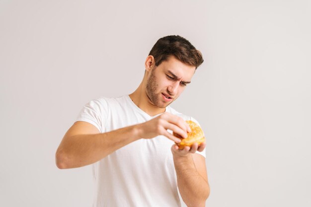 Porträt eines unzufriedenen jungen Mannes mit Ekel, der die Knospe eines schlechten Burgers auf weißem, isoliertem Hintergrund pflückt. Studioaufnahme eines gutaussehenden bärtigen Mannes, der ungesunde, köstliche Hamburger in den Händen hält.