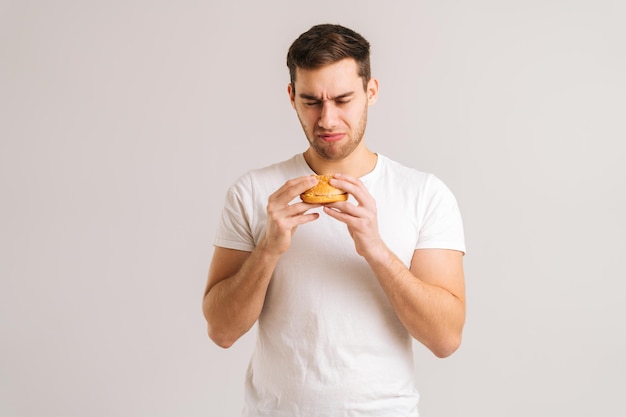 Porträt eines unzufriedenen jungen Mannes mit Ekel, der einen schlechten Burger auf weißem, isoliertem Hintergrund isst. Studioaufnahme eines gutaussehenden bärtigen Mannes, der einen ungesunden Cheeseburger in den Händen hält.