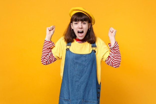 Porträt eines verängstigten, schreienden Mädchen-Teenagers in französischer Baskenmütze, Denim-Sommerkleid, das die Fäuste isoliert auf gelbem Wandhintergrund im Studio ballt. Menschen aufrichtige Emotionen, Lifestyle-Konzept. Mock-up-Kopienbereich.