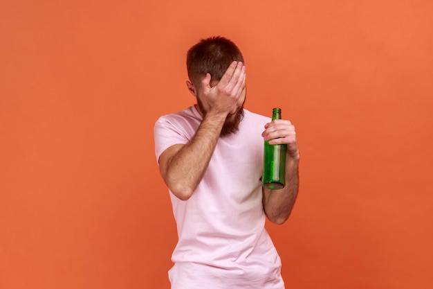 Porträt eines verärgerten traurigen bärtigen Mannes, der mit einer Bierflasche steht, betrunken ist, sein Gesicht mit einer Handfläche bedeckt und ein rosa T-Shirt trägt. Studioaufnahme im Innenbereich isoliert auf orangefarbenem Hintergrund.