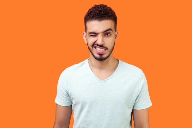Porträt eines verspielten, gutaussehenden brünetten Mannes mit Bart im lässigen weißen T-Shirt, der mit einem geschlossenen Auge steht, zwinkert und mit einem Grinsen in die Kamera flirtet, Studioaufnahme isoliert auf orangefarbenem Hintergrund