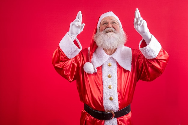 Porträt eines verträumten älteren Mannes im Weihnachtsmann-Kostüm, das die Finger kreuzt, um einen Wunsch zu machen, Weihnachtszauber, Winterferien. Indoor-Studioaufnahme auf rotem Hintergrund isoliert