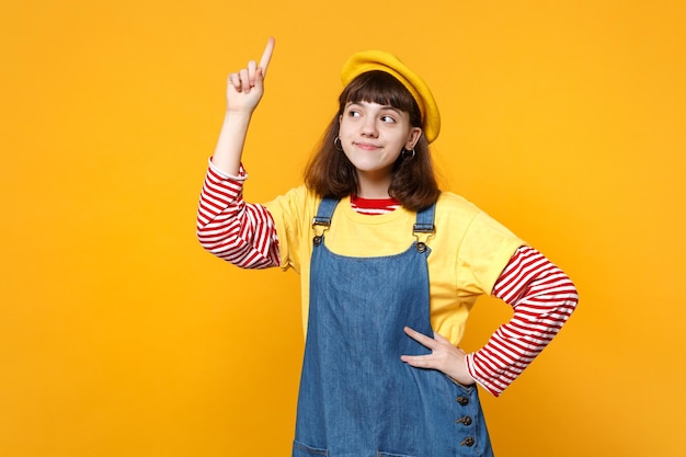 Porträt eines verträumten Mädchen-Teenagers in französischer Baskenmütze, Denim-Sommerkleid, das den Zeigefinger nach oben zeigt, isoliert auf gelbem Wandhintergrund im Studio. Menschen aufrichtige Emotionen Lifestyle-Konzept. Mock-up-Kopienbereich.