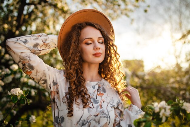 Porträt eines verträumten Mädchens am Frühlingspark unter blühenden weißen Bäumen auf einem Sonnenuntergang. Frau, die beige Hut und weißes Kleid trägt, riecht Blüte der Apfelbäume. Frühlingssaison-Konzept.