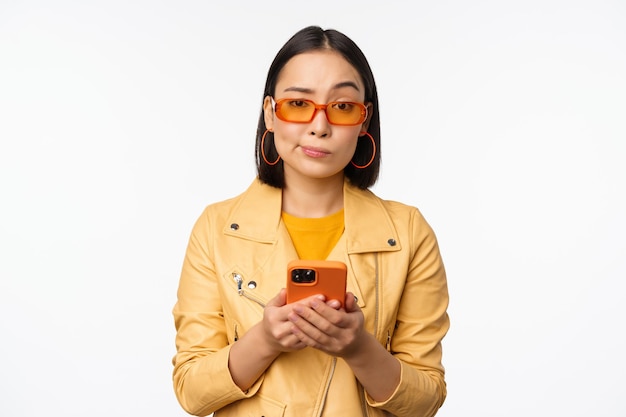 Porträt eines verwirrten asiatischen Mädchens mit Sonnenbrille, das ein Smartphone mit einem Handy hält und verwirrt aussieht, wie es ahnungslos vor weißem Hintergrund steht