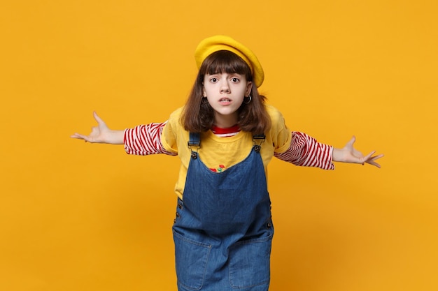 Porträt eines verwirrten, gereizten Teenagers in französischer Baskenmütze, Denim-Sommerkleid, das die Hände isoliert auf gelbem Hintergrund im Studio ausbreitet. Menschen aufrichtige Emotionen, Lifestyle-Konzept. Mock-up-Kopienbereich.