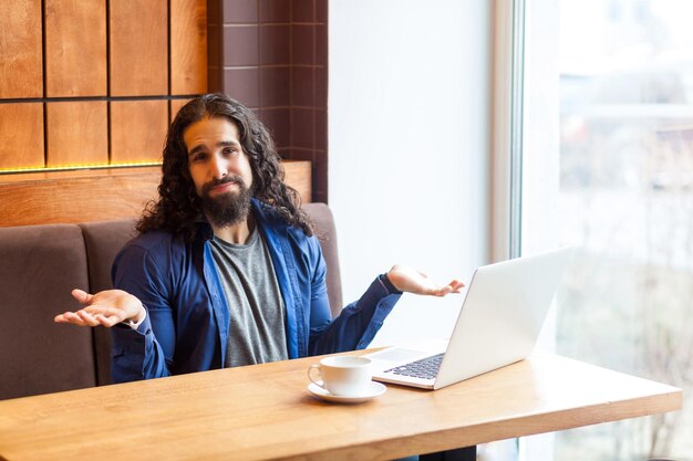 Porträt eines verwirrten, gutaussehenden jungen erwachsenen Freiberuflers im lässigen Stil, der im Café mit Laptop sitzt, die Hände erhebt und eine unbekannte Geste zeigt, in die Kamera schaut. Indoor, Lifestyle-Konzept