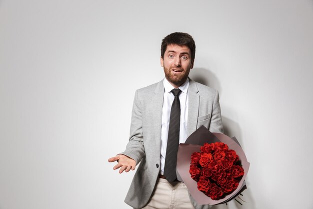 Porträt eines verwirrten jungen Mannes im Anzug, der isoliert steht und Strauß von Rosen hält