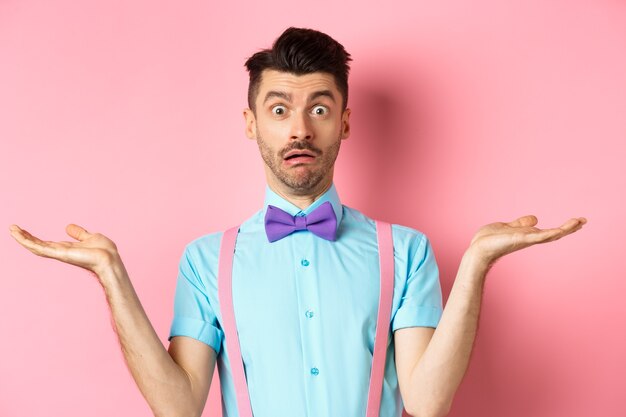Porträt eines verwirrten männlichen Models in Fliege und lustigem Hemd, achselzuckend und ahnungslos, weiß nichts, steht auf rosafarbenem Hintergrund.