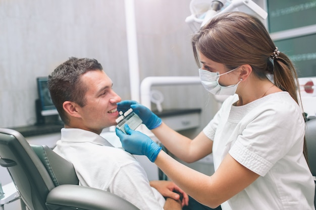 Porträt eines weiblichen Zahnarztes und des jungen glücklichen männlichen Patienten in einem Zahnarztbüro. Mann, der Farbe der Zähne überprüft und vorwählt