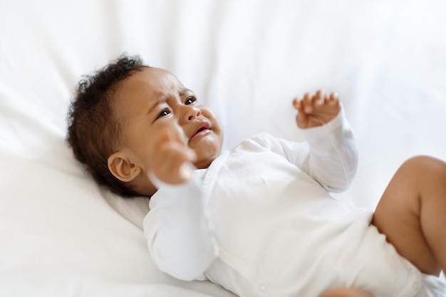 Porträt eines weinenden kleinen schwarzen Babys, das einen Body trägt, der auf dem Bett liegt