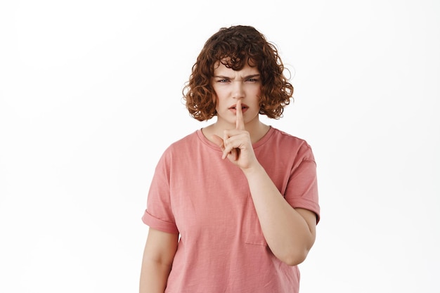 Porträt eines wütenden kaukasischen Mädchens, sagen, ruhig zu sein, enttäuscht die Stirn runzeln und schweigen, den Finger in der Tabu-Geste drücken, nicht sprechen, im T-Shirt vor weißem Hintergrund stehen.