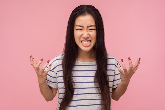 Porträt eines wütenden, wütenden Mädchens mit langen Haaren in gestreiftem T-Shirt, das mit zusammengebissenen Zähnen und erhobenen Händen steht und wilde Wut ausdrückt, die sich verrückt fühlt, Studioaufnahme isoliert auf rosa Hintergrund