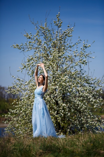 Porträt eines zarten Mädchens in einem blauen langen Kleid mit Tüll unter blühenden Kirschen mit einem Lächeln im Gesicht an einem sonnigen warmen Sommertag