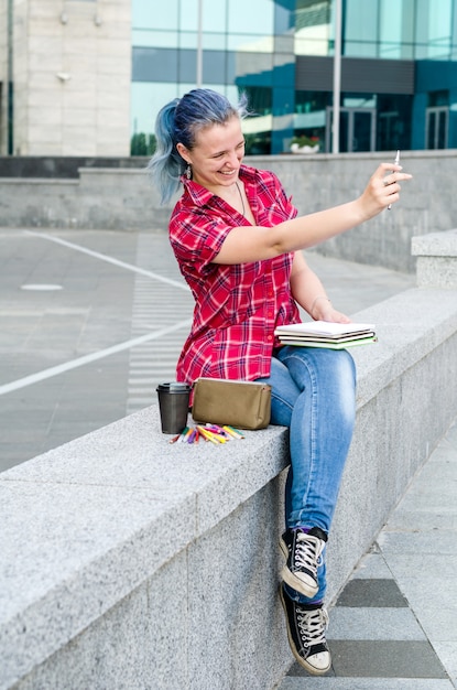 Porträt eines zufälligen netten und jungen Mädchens mit dem blauen Haar in den Jeans in der städtischen Sommerzeichnung oder im Schreiben etwas in ein Notizbuch