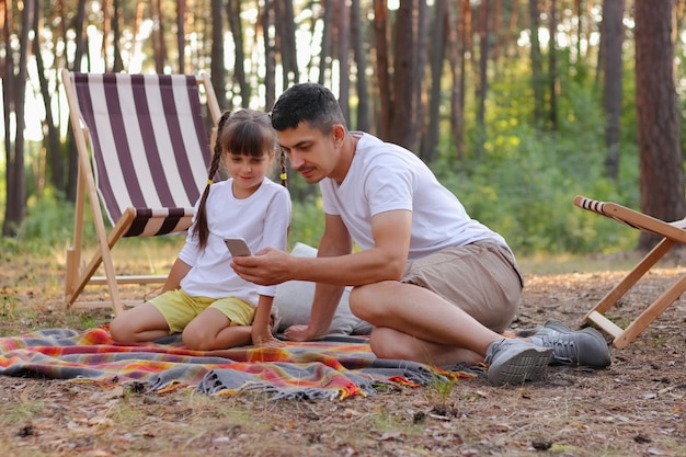 Porträt eines zufriedenen Familienvaters und einer Tochter, die im Parkwald um Bäume herum sitzen. Kleine Mädchen, Tochter und ihr Vater, die sich mit dem Handy Fotos von ihrer Ruhe ansehen und Spaß haben