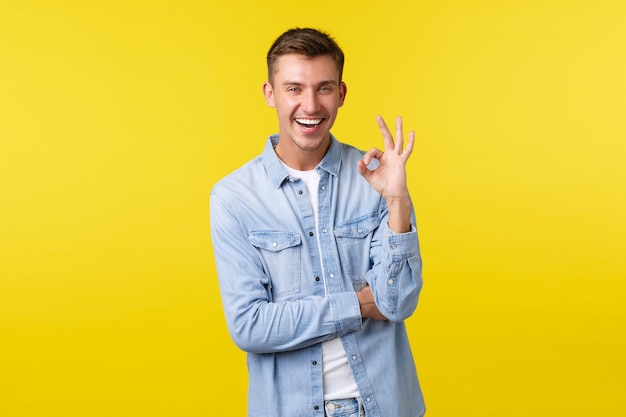 Porträt eines zufriedenen, gutaussehenden, glücklichen männlichen Studenten, Kerl, der eine zustimmende Geste zeigt. Mann macht OK-Zeichen und lacht sorglos, genießt tolle Sommer-Promo-Angebote, gelber Hintergrund.