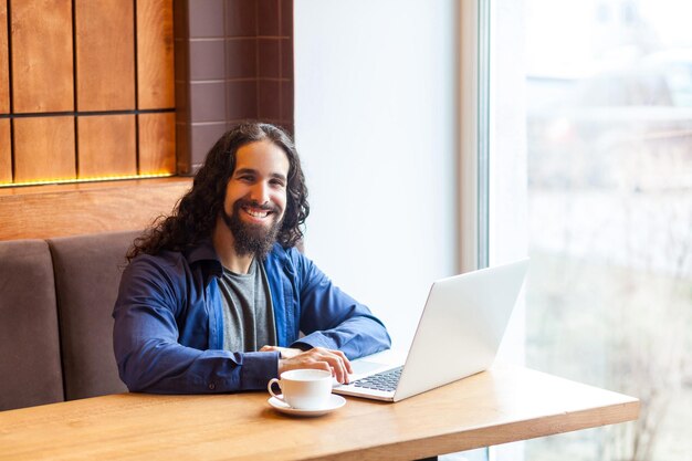 Porträt eines zufriedenen, gutaussehenden, intelligenten, bärtigen jungen erwachsenen Mannes, Freiberufler im lässigen Stil, der im Café sitzt und am Laptop arbeitet, in die Kamera schaut, Geschäftsmann im Büro. Indoor, Lifestyle-Konzept