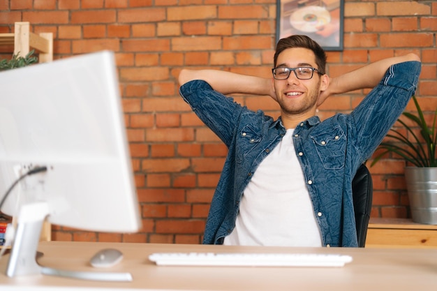 Porträt eines zufriedenen, gutaussehenden jungen freiberuflichen Programmierers mit stylischer Brille, der die Hände hinter dem Kopf hält und am Schreibtisch mit Desktop-Computer sitzt