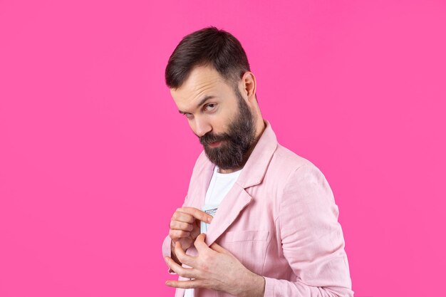 Porträt eines zufriedenen jungen Geschäftsmannes mit Bart, gekleidet in eine rosa Jacke, die US-Dollar-Banknoten vor einem roten Studiohintergrund zeigt Schmecken Sie den Geruch von Geld
