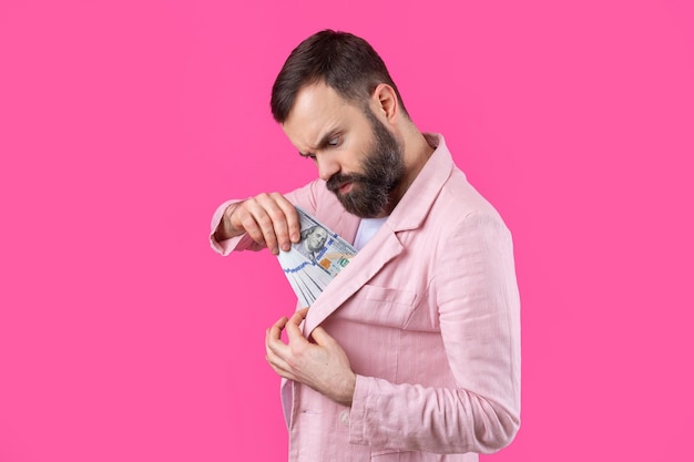 Porträt eines zufriedenen jungen Geschäftsmannes mit Bart in einer rosa Jacke, die US-Dollar-Banknoten vor einem roten Studiohintergrund zeigt Schmecken Sie den Geruch von Geld