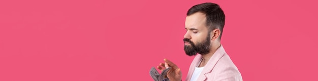 Porträt eines zufriedenen jungen Geschäftsmannes mit Bart in einer rosa Jacke, die US-Dollar-Banknoten vor einem roten Studiohintergrund zeigt Schmecken Sie den Geruch von Geld