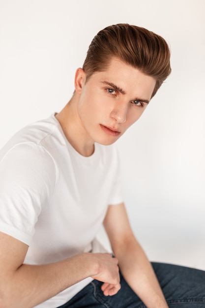 Porträt Europäischer attraktiver junger Mann mit stilvoller Frisur mit sauberer Haut in modischem, klassischem weißem T-Shirt in blauen Vintage-Jeans in der Nähe der Vintage-Wand im Studio. Hübscher Kerl ruht sich drinnen auf Stuhl aus