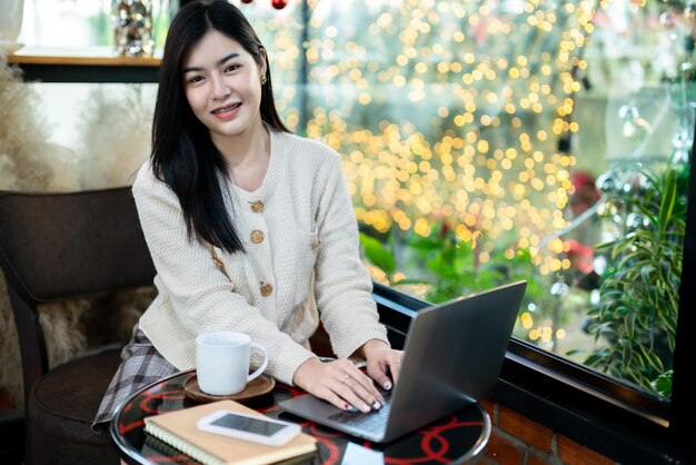 Porträt freiberuflich tätiges schönes positives Lächeln junge asiatische Frau, die online mit Laptop-Computer und Smartphone auf Notebook zu Hause im Wohnzimmer drinnen oder im CaféBusiness-Lifestyle arbeitet