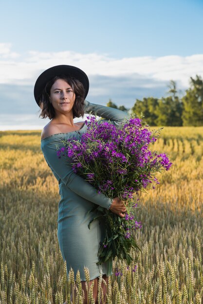 Porträt freudige junge Frau Brünette im blauen Kleid mit einem Strauß lila Blumen, die auf der schönen Natur auf Feld gehen und ruhen. Stilvolle Hipster-Frau. Atmosphärisches Lifestyle-Foto im Freien