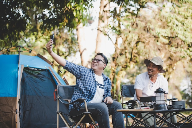 Porträt glücklicher asiatischer Mann Freunde Einen Videoanruf mit Smartphone auf dem Campingplatz machen Kochset Vorderboden Kochen im Freien Reisen Camping Lifestyle-Konzept