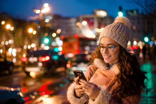 Porträt im Freien der schönen jungen Frau, die ihren Handy nachts nahe Stadtstraße verwendet.