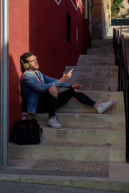 Porträt im Freien des modernen jungen Mannes mit dem intelligenten Telefon, das in der Straße sitzt.