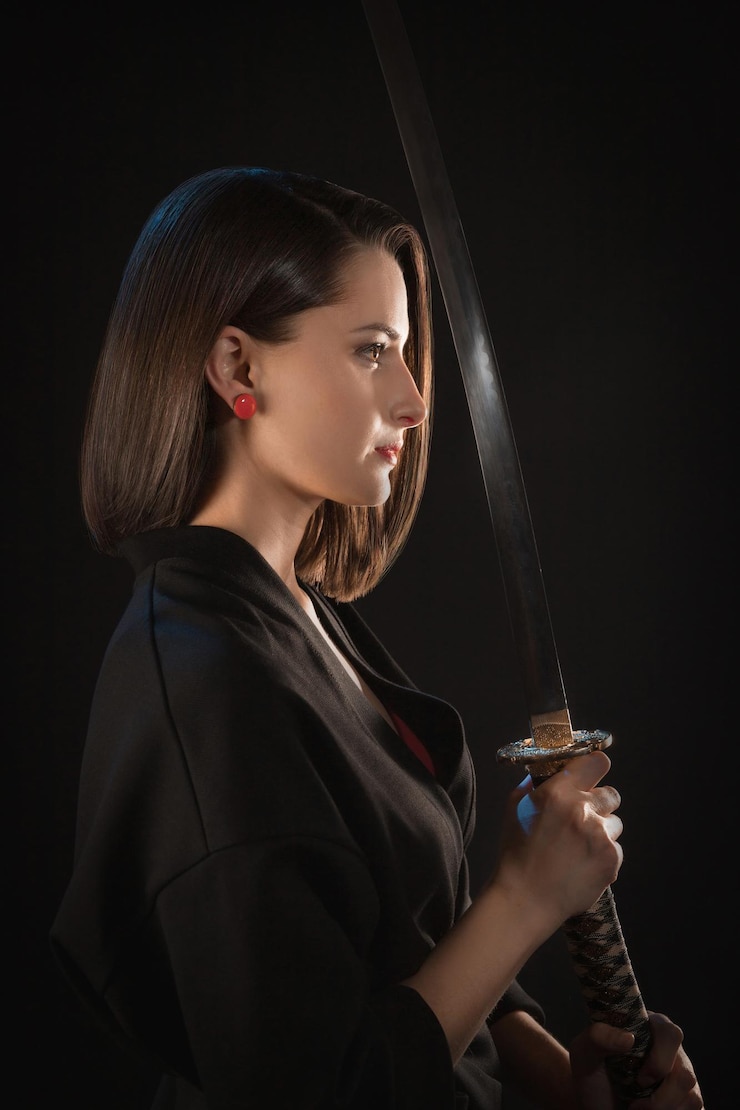 Porträt im Profil einer jungen Frau, die ein Katana in ihren Händen vor sich auf schwarzem Hintergrund hält