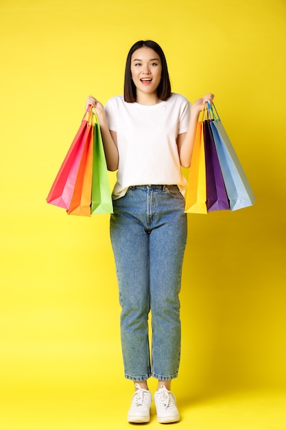 Porträt in voller Größe des schönen asiatischen Mädchens, das einkaufen geht, Papiertüten von Geschäften hält und lächelt, in Jeans und weißem T-Shirt über gelbem Hintergrund stehend.