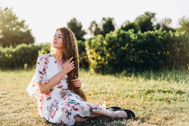 Porträt in voller Länge der schönen Frau mit dem langen lockigen Haar auf Johannisbeerfeldhintergrund. Mädchen in einem hellen Kleid sitzt auf dem Gras im sonnigen Tag