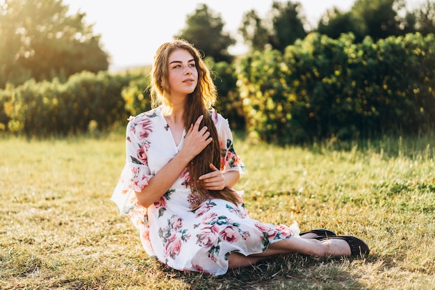 Porträt in voller Länge der schönen Frau mit dem langen lockigen Haar auf Johannisbeerfeldhintergrund. Mädchen in einem hellen Kleid sitzt auf dem Gras im sonnigen Tag