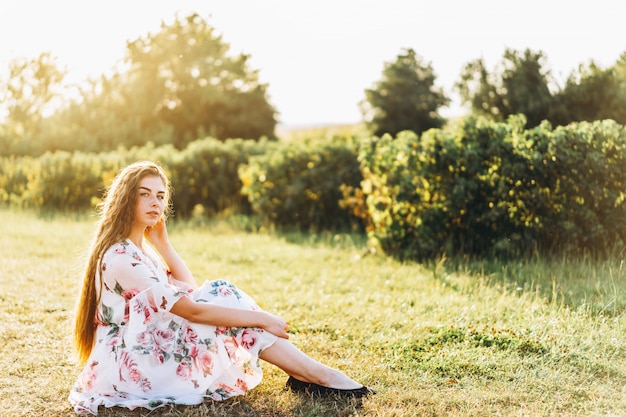 Porträt in voller Länge der schönen Frau mit dem langen lockigen Haar auf Johannisbeerfeldhintergrund. Mädchen in einem hellen Kleid sitzt auf dem Gras im sonnigen Tag