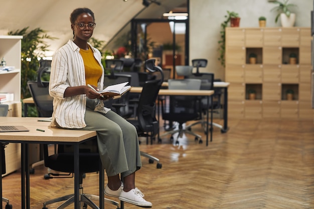 Porträt in voller Länge der zeitgenössischen afroamerikanischen Frau, die auf Schreibtisch im Büro sitzt und selbstbewusst Raum kopiert