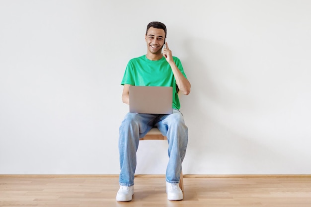 Porträt in voller Länge des Kerls mit dem Laptop, der auf dem Handy sitzt auf Stuhl gegen weißen Wandkopienraum kommuniziert