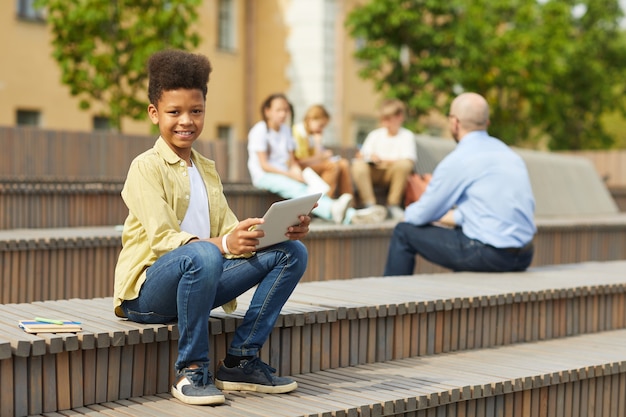 Porträt in voller Länge des lächelnden afroamerikanischen Jungen, der Kamera beim Sitzen auf Bank im Freien mit Lehrer betrachtet, der Lektion im Hintergrund, Kopienraum gibt