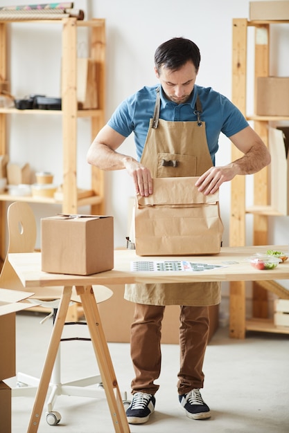 Porträt in voller Länge des reifen Mannes, der Schürzenverpackungsaufträge trägt, während er durch Holztisch, Lebensmittel-Lieferservice-Arbeiter steht