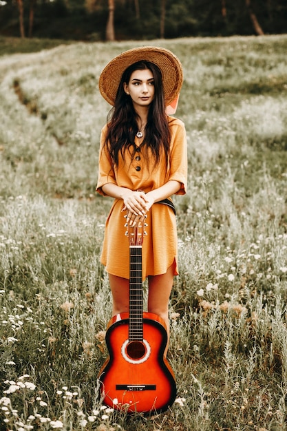 Porträt in voller Länge einer erstaunlichen Frau mit dunklen langen Haaren, die wegschauen, während sie sich auf eine Gitarre gegen einen Hügel mit Blumen lehnen.