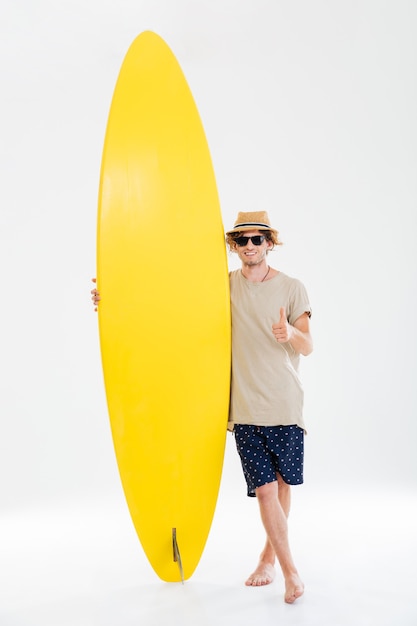 Porträt in voller Länge eines lächelnden glücklichen Mannes in der Sonnenbrille und im Hut, die Daumen hoch Geste zeigen und Surfbrett lokalisiert auf der weißen Wand halten