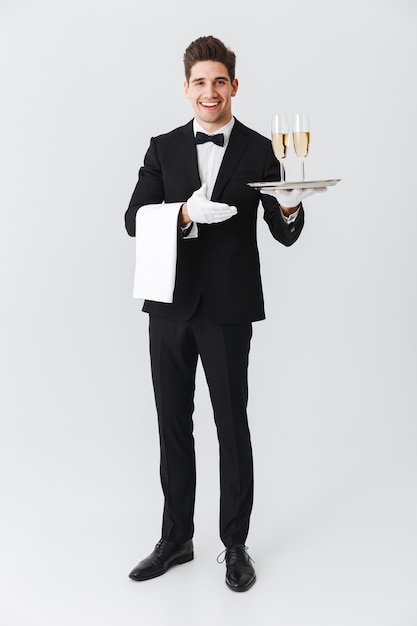 Porträt in voller Länge eines lächelnden jungen Kellners im Smoking, der Tablett mit zwei Gläsern Champagner über weißer Wand hält
