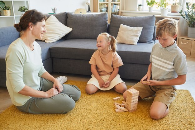 Porträt in voller Länge von liebevoller Familie mit Kindern mit besonderen Bedürfnissen, die Brettspiele spielen, während sie zu Hause auf dem Boden sitzen, Platz kopieren