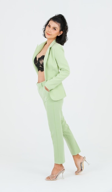 Porträt isolierte Ausschnitt-Studioaufnahme einer asiatischen sexy Modegeschäftsfrau mit lockigem Haar in grünem Anzug mit schwarzem Spitzen-Dessous-Crop-Top-BH, der stehend posiert und die Hände in die Hosentaschen auf weißem Hintergrund steckt