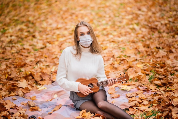 Porträt junge Frau in der Schutzmaske, die Ukulele-Gitarre im Herbstpark spielt