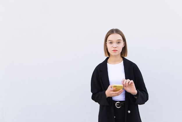 Porträt junge Geschäftsfrau, die mit einem Smartphone in ihren Händen auf weißer Wand steht